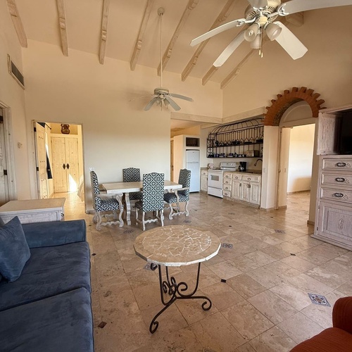 La imagen muestra un amplio espacio interior de planta abierta que integra sala, comedor y cocina, con techos altos de vigas vistas, ventiladores de techo y suelos de baldosas decoradas.