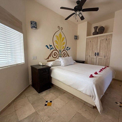 Un dormitorio con una cama blanca decorada con pétalos de rosa, un mural colorido en la pared, suelo de baldosas y un ventilador de techo.