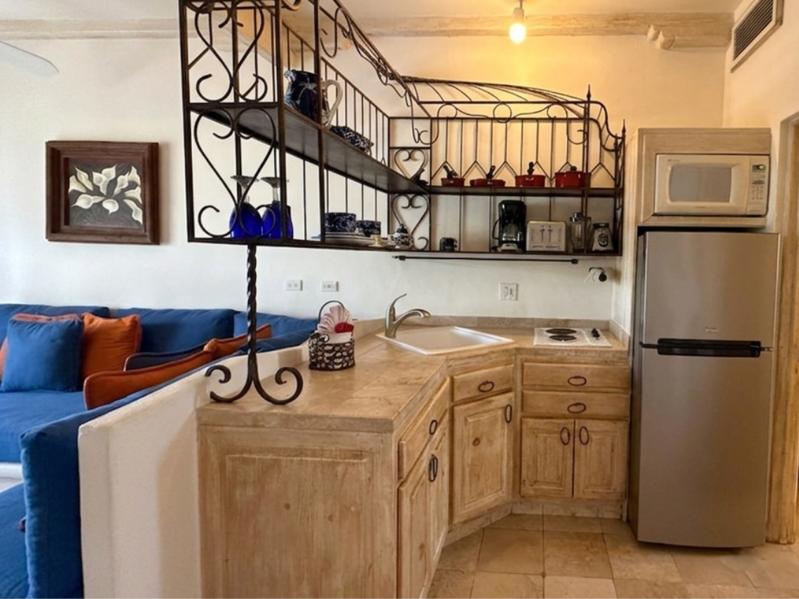 A cozy, open-plan studio apartment features a blue sofa, a kitchenette with wooden cabinets and wrought iron shelving, and a beamed ceiling, all in a warm, Southwestern-inspired style.