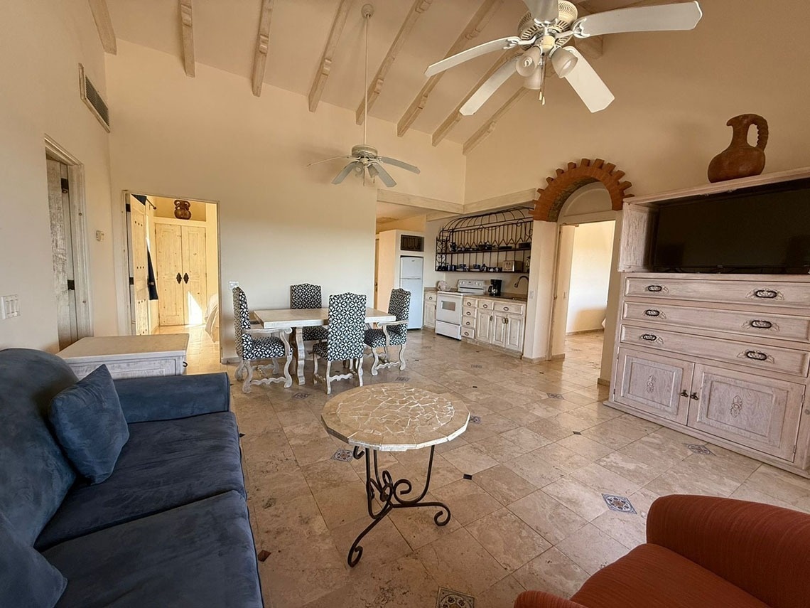 Un salón-comedor de estilo rústico con cocina abierta, muebles de madera clara, suelo de baldosas y techos con vigas, donde destacan un sofá azul y un gran mueble con televisión.
