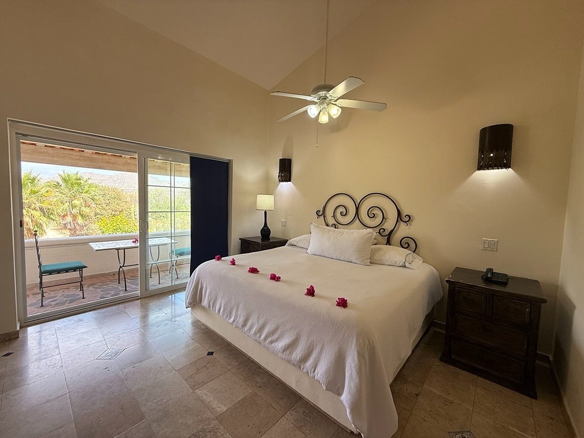 Un dormitorio luminoso con una cama blanca adornada con flores rosadas, un ventilador de techo y un balcón con vistas a palmeras y montañas.