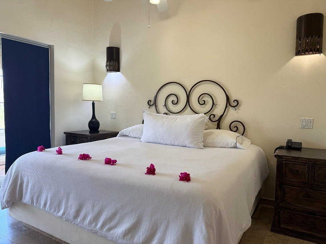 Un dormitorio luminoso presenta una cama grande con cabecero de hierro forjado, adornada con flores rosas, flanqueada por mesitas de noche y apliques de pared.