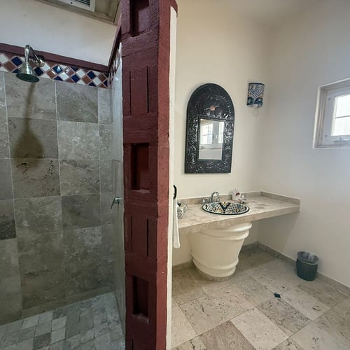 Un baño de estilo tradicional con ducha de azulejos grises y un lavabo de pedestal con tazón decorado, separados por una estructura divisoria de ladrillo rojo.