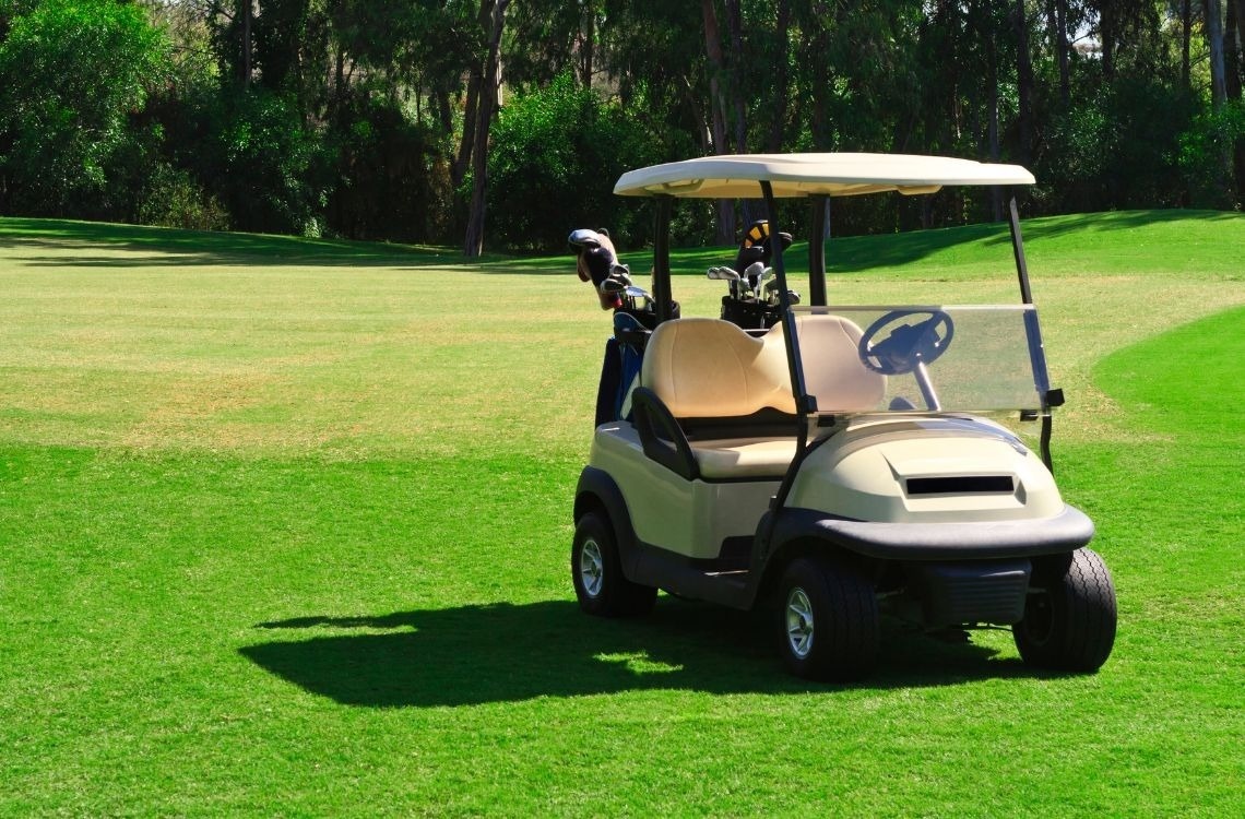A golf cart with golf clubs is parked on a well-maintained green golf course, with trees lining the background under sunny conditions.