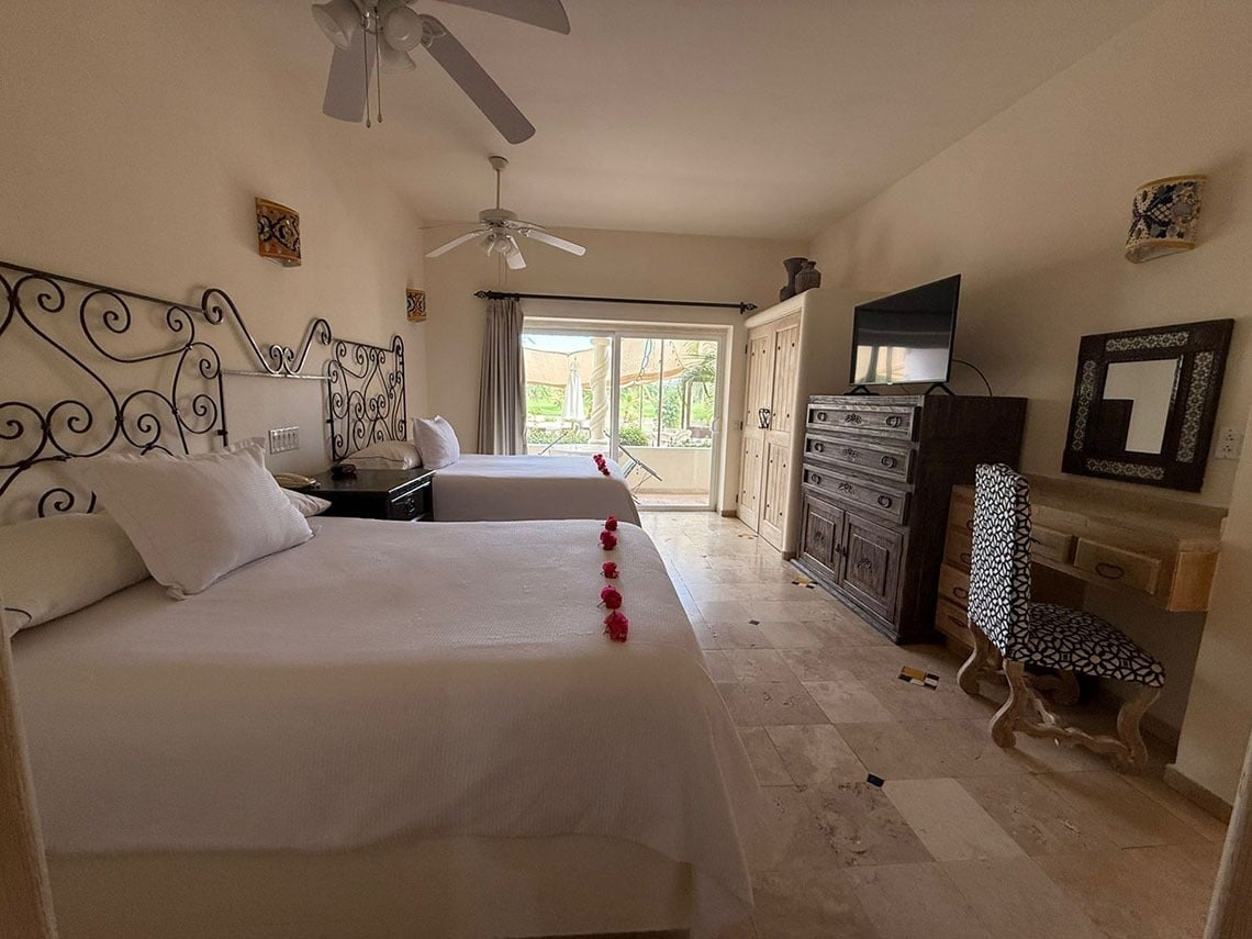 Un luminoso dormitorio con dos camas decoradas con pétalos rojos, ventiladores de techo y una zona de escritorio y televisión, con acceso a una terraza exterior.