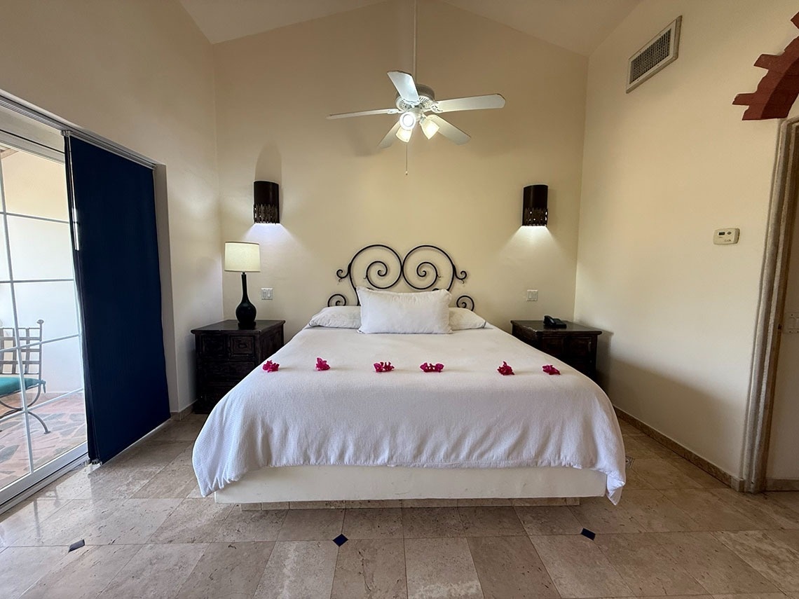 Un dormitorio luminoso con una cama blanca decorada con pétalos rojos, mesitas de noche, ventilador de techo y una puerta corrediza de vidrio que da a un patio.