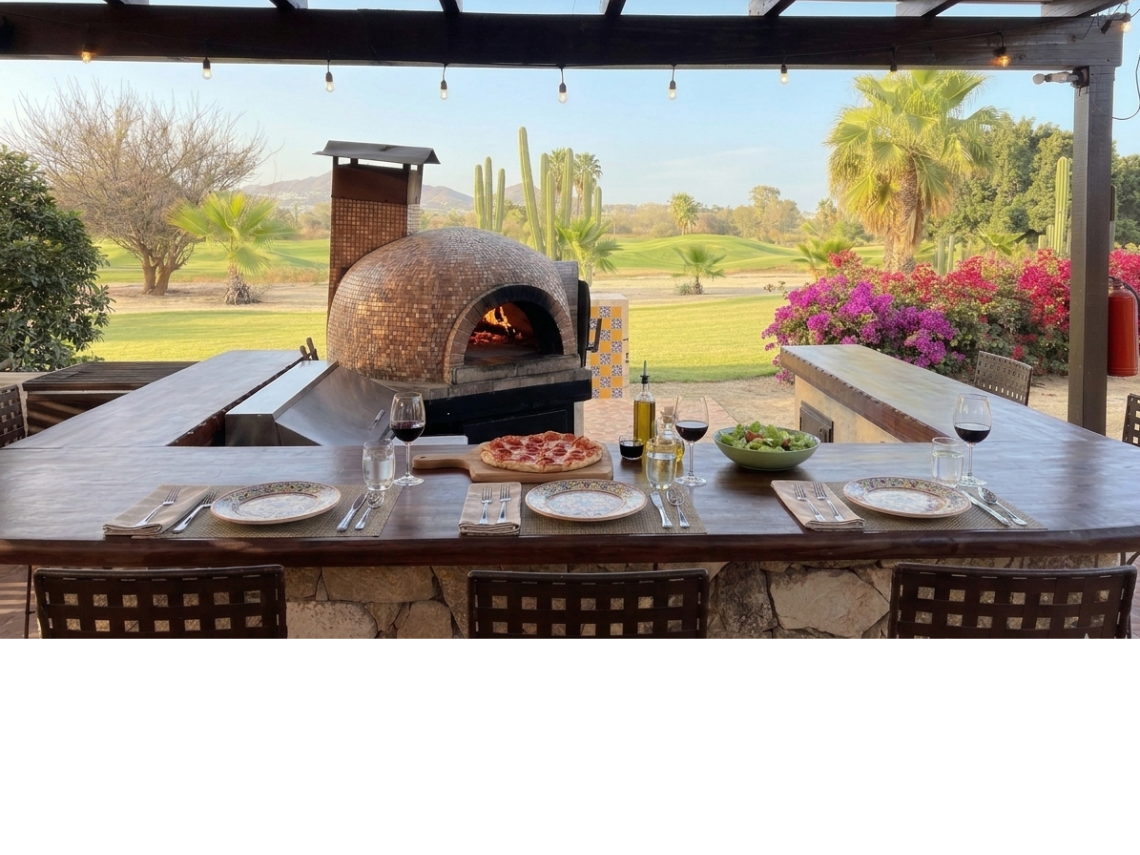 This image captures a spacious outdoor patio area, featuring shaded seating, a bar with a pizza oven, and a desert landscape with cactuses and distant hills under a partly cloudy sky.