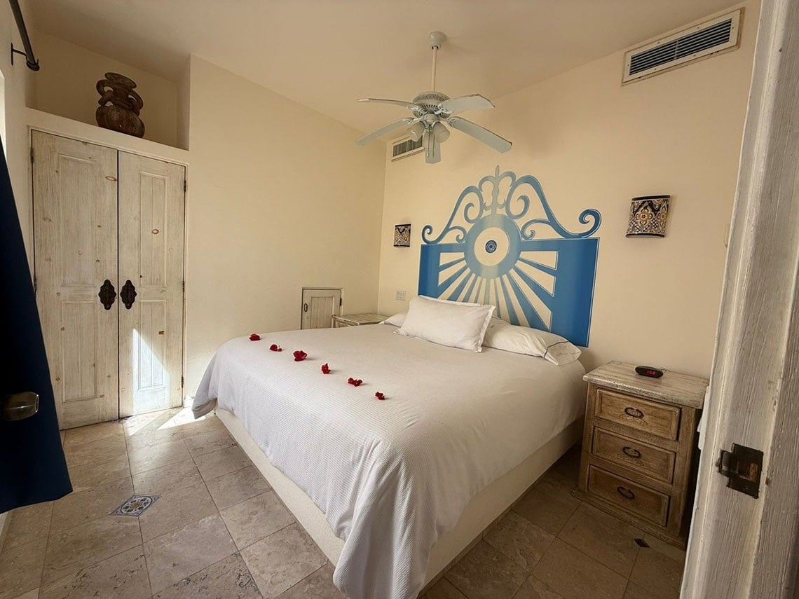 Una habitación luminosa y acogedora con una cama blanca decorada con pétalos de rosa, un cabecero azul pintado y suelo de baldosas.