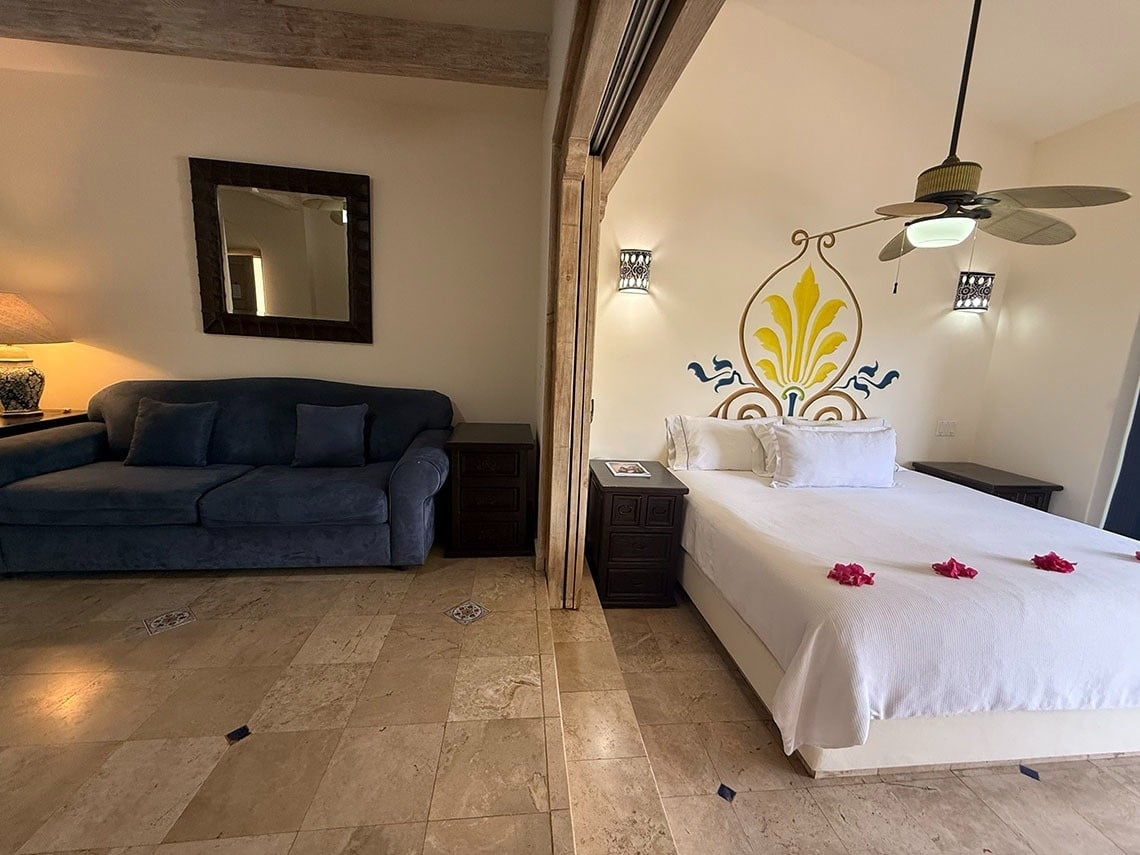 Una suite de hotel presenta un área de sala de estar con un sofá azul y un espejo, y un dormitorio con una cama blanca adornada con pétalos rojos y un cabecero pintado, bajo un ventilador de techo.