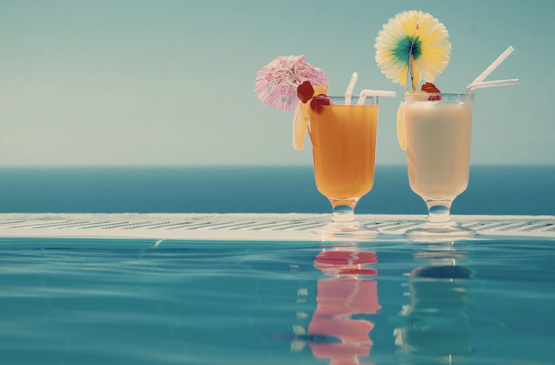 Una piscina de resort con tumbonas y sombrillas bajo el sol está rodeada de edificios de estilo mediterráneo o colonial.