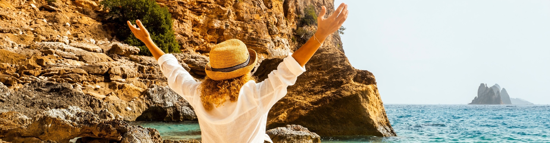 Joyful woman on beach with arms up, enjoying sea view and rocky cliffs.
