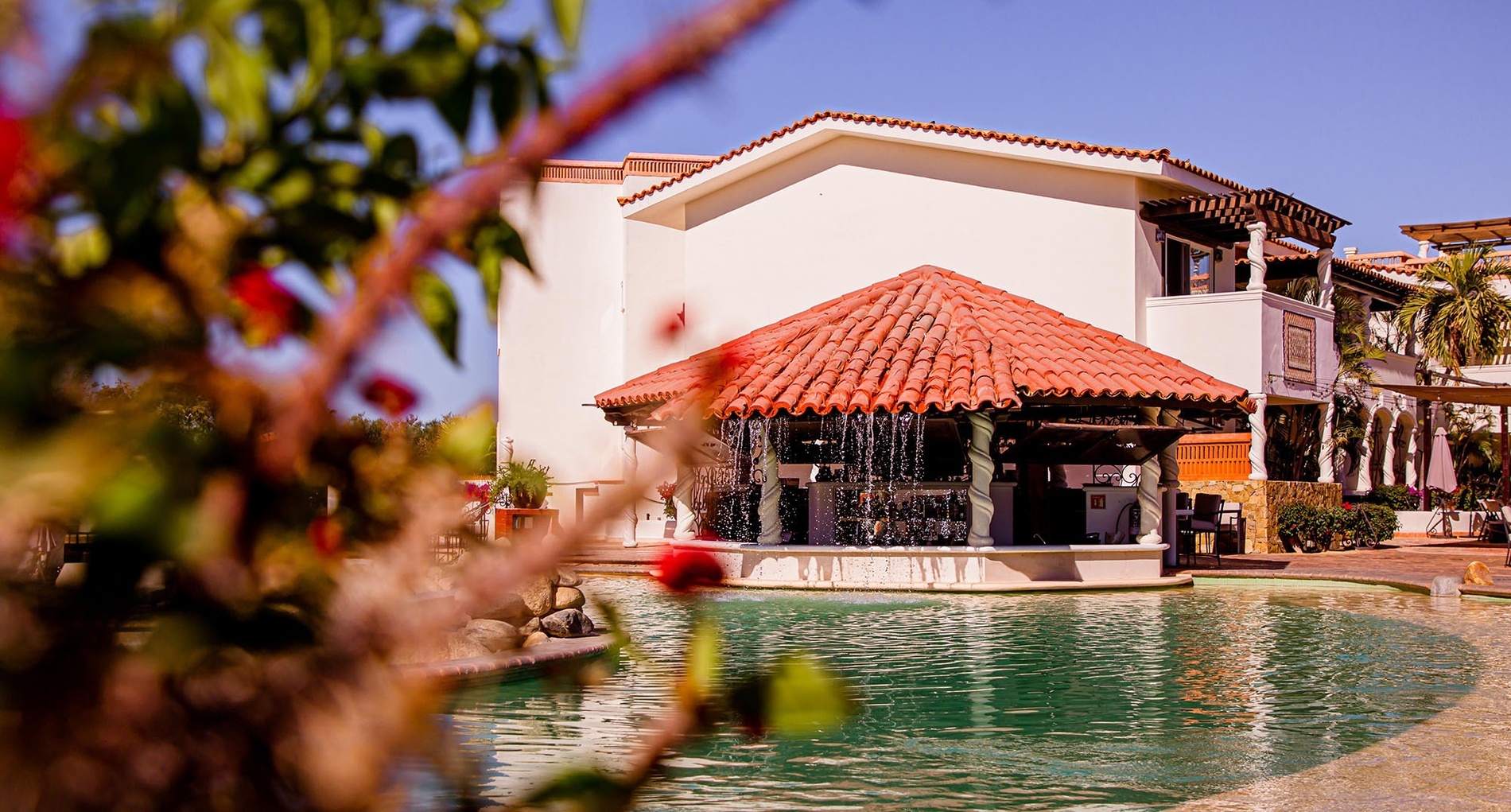 Un bar con techo de tejas rojas y cascadas de agua dentro de una piscina, flanqueado por un edificio blanco y follaje borroso, se ve bajo un cielo azul.