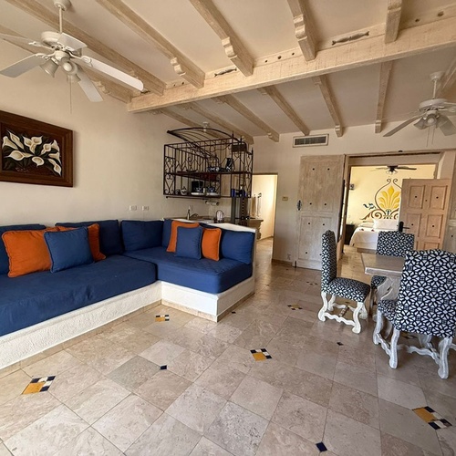 Una luminosa sala de estar con un sofá en forma de L azul y cojines naranjas, una mesa de comedor con sillas estampadas y vigas de madera expuestas en el techo, abierta a un dormitorio adyacente.