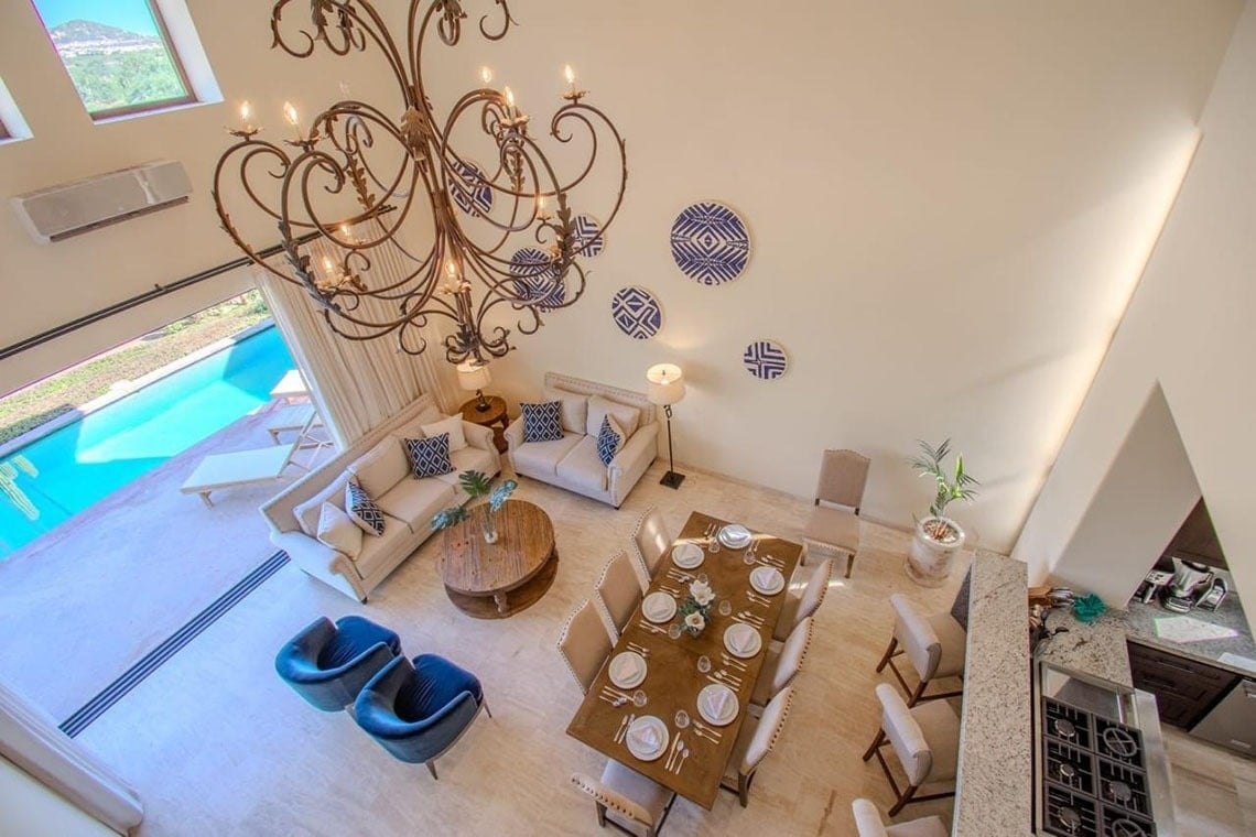Una vista aérea muestra un espacioso salón de planta abierta con un gran candelabro, áreas de comedor y sala de estar elegantemente decoradas, y una piscina visible a través de grandes ventanales.