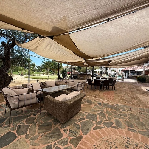 La imagen muestra un amplio patio exterior sombreado por lonas, equipado con sofás y mesas, y con un campo de golf de fondo.