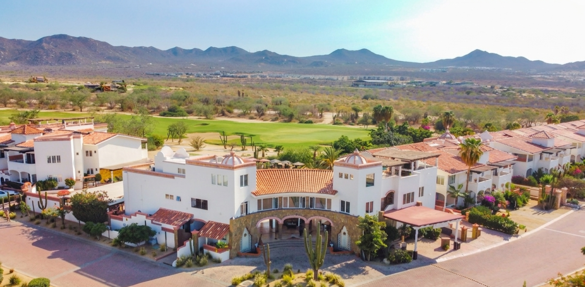 An aerial view captures the white buildings of the Los Cabos Golf Resort and its green fairways nestled within a desert landscape, with mountains in the distant background.