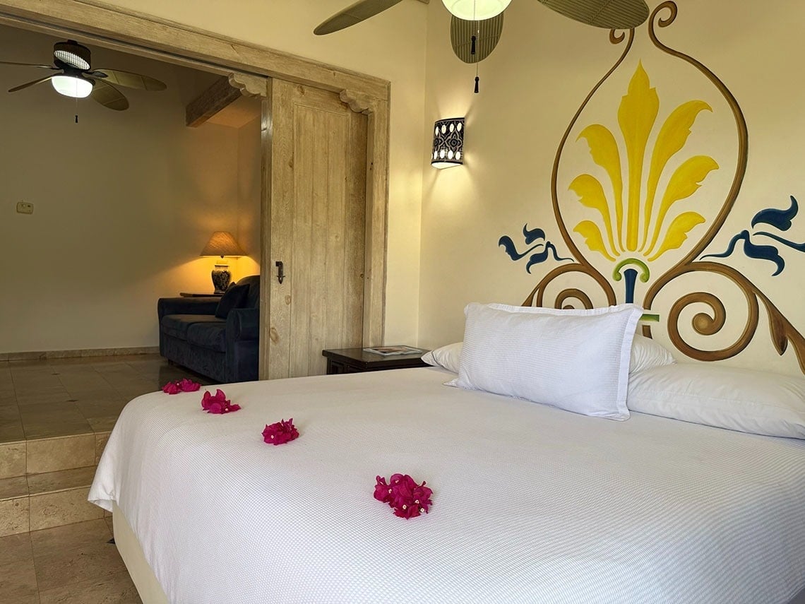 Una suite de hotel decorada con una cama blanca con flores rosadas, un mural vibrante en la pared y una puerta corredera de madera hacia una sala de estar.