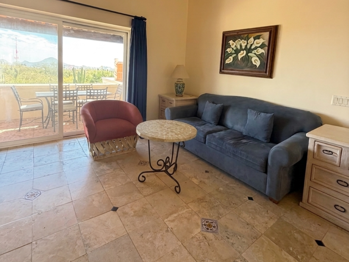 An interior living room features a blue sofa, a red armchair, a central coffee table, and a ceiling fan, with a large sliding glass door opening to a balcony offering a scenic view of mountains and vegetation.