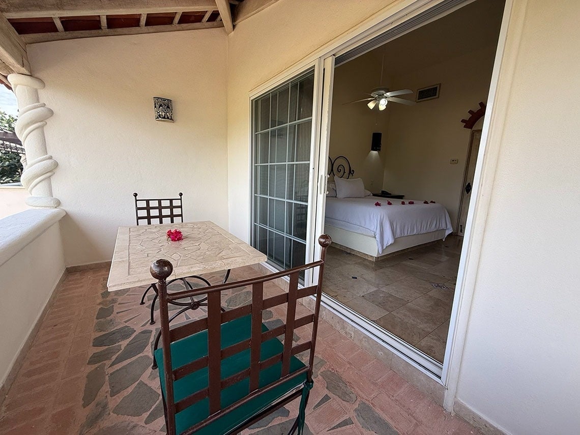 Una terraza exterior con una mesa y sillas se conecta a un dormitorio con una cama decorada con pétalos rojos a través de una puerta corrediza de vidrio.