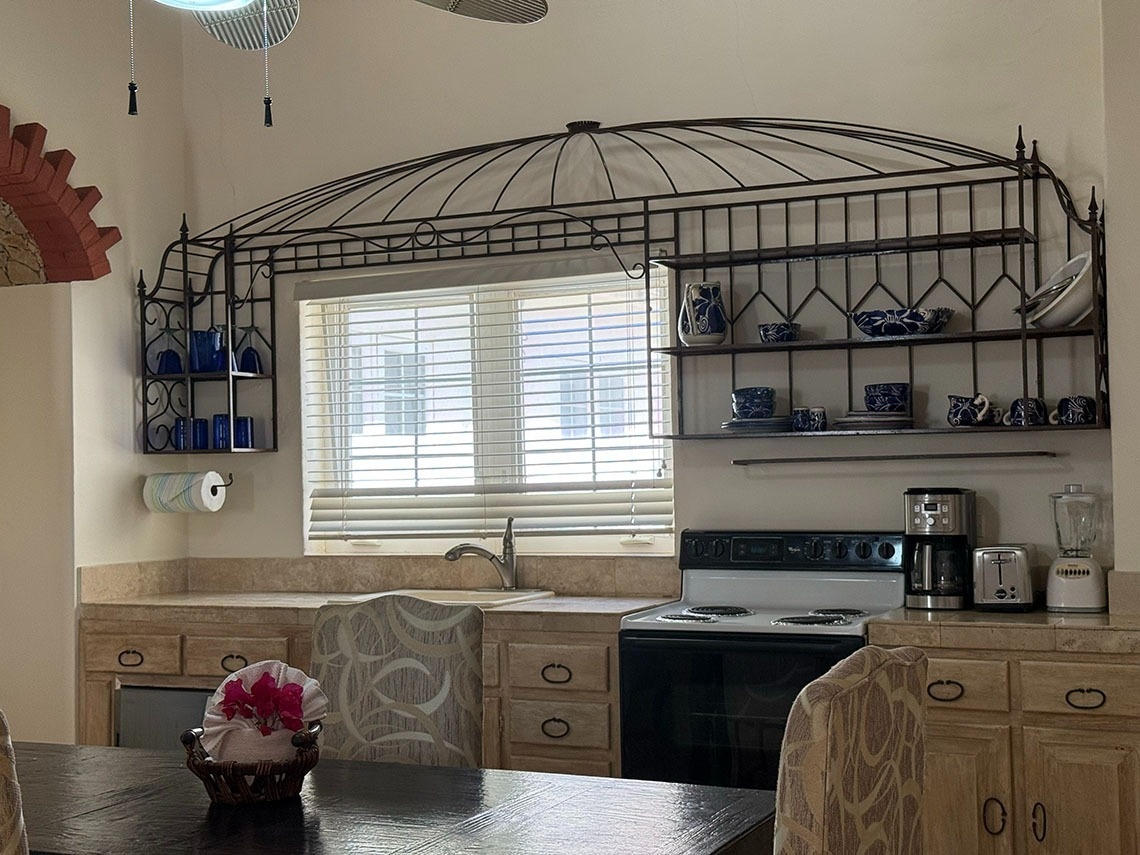 Una cocina con una ventana, estantes decorativos de metal exhibiendo vajilla azul y blanca, electrodomésticos de cocina y una mesa de comedor con un centro de flores en primer plano.