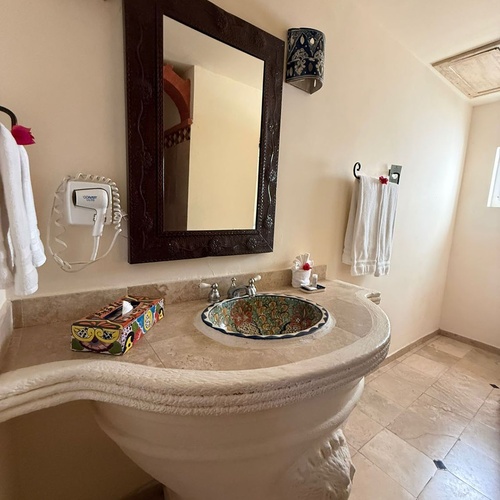 Un baño luminoso y ricamente decorado presenta un tocador de pedestal esculpido con un vibrante lavabo de cerámica pintado a mano y un gran espejo enmarcado.