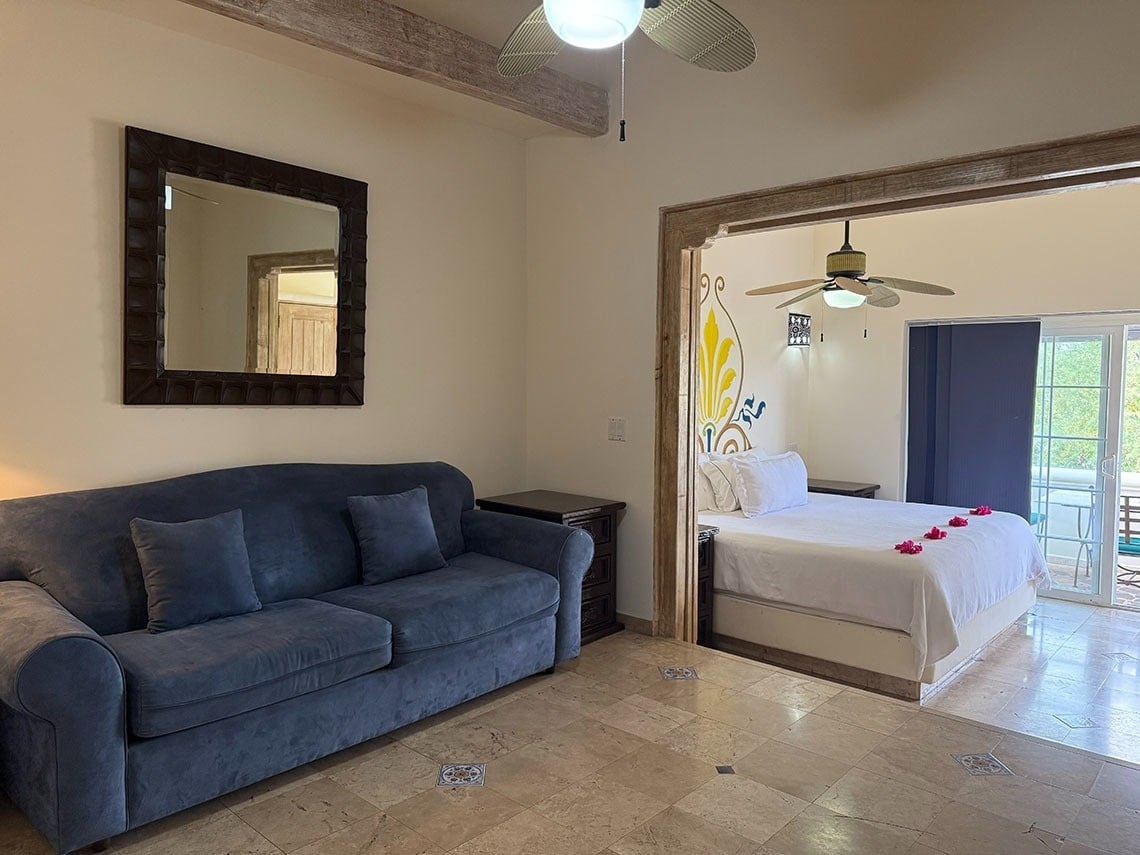 Una sala de estar con un sofá azul y un espejo grande se conecta a un dormitorio con una cama blanca y un balcón a través de una abertura arqueada, ambos con suelos de baldosas y ventiladores de techo.