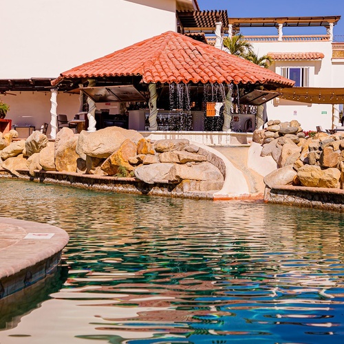 Un bar de piscina con tejado rojo y cascadas de agua se erige frente a un edificio de resort, con las aguas azules de la piscina en primer plano bajo un cielo claro.