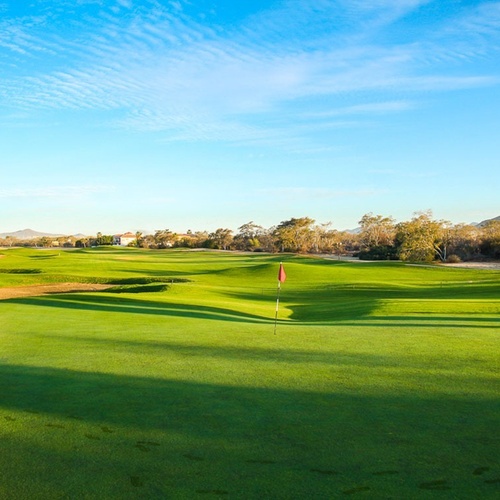 Un exuberante campo de golf verde con una bandera roja se extiende bajo un cielo azul vibrante con nubes ligeras, flanqueado por árboles y montañas distantes.
