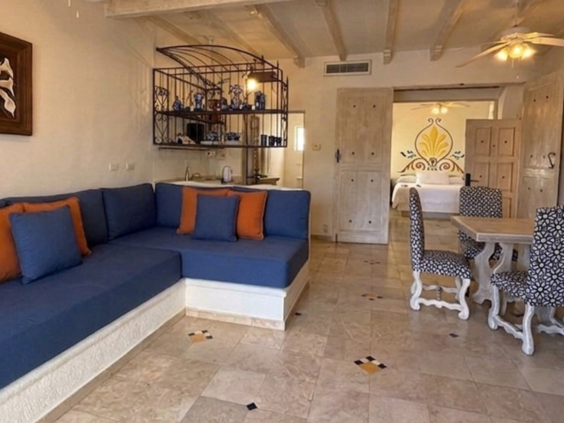 This inviting living space features a blue L-shaped sofa, a dining area with patterned chairs, rustic wooden ceiling beams, and a tiled floor, with an open doorway leading to a bedroom.