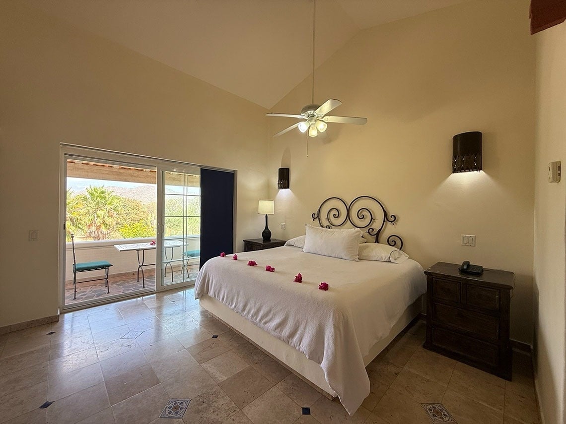 Dormitorio con cabecero de hierro forjado, suelo de baldosas y balcón con vistas a un paisaje natural exuberante.