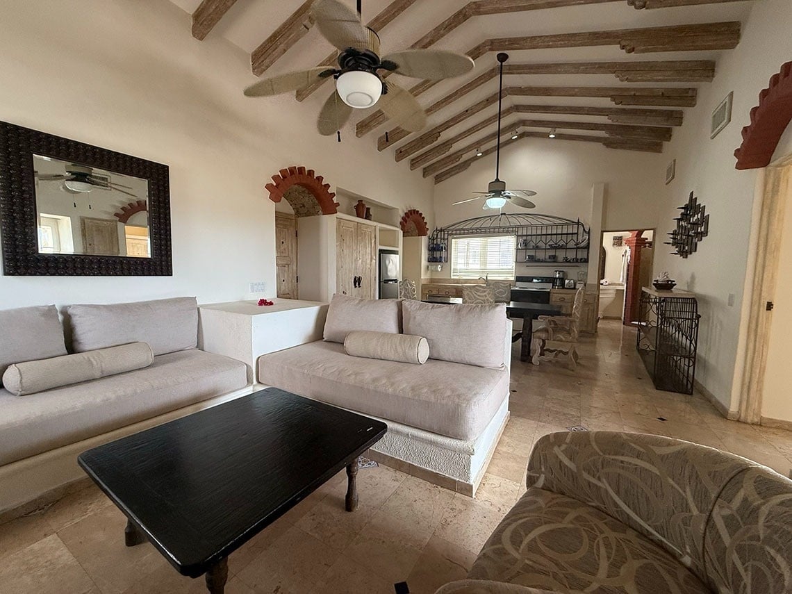 Amplia sala de estar de estilo rústico-mediterráneo con vigas de madera expuestas en el techo, sofás claros y arcos de ladrillo, que se conecta a un área de cocina y comedor en el fondo.