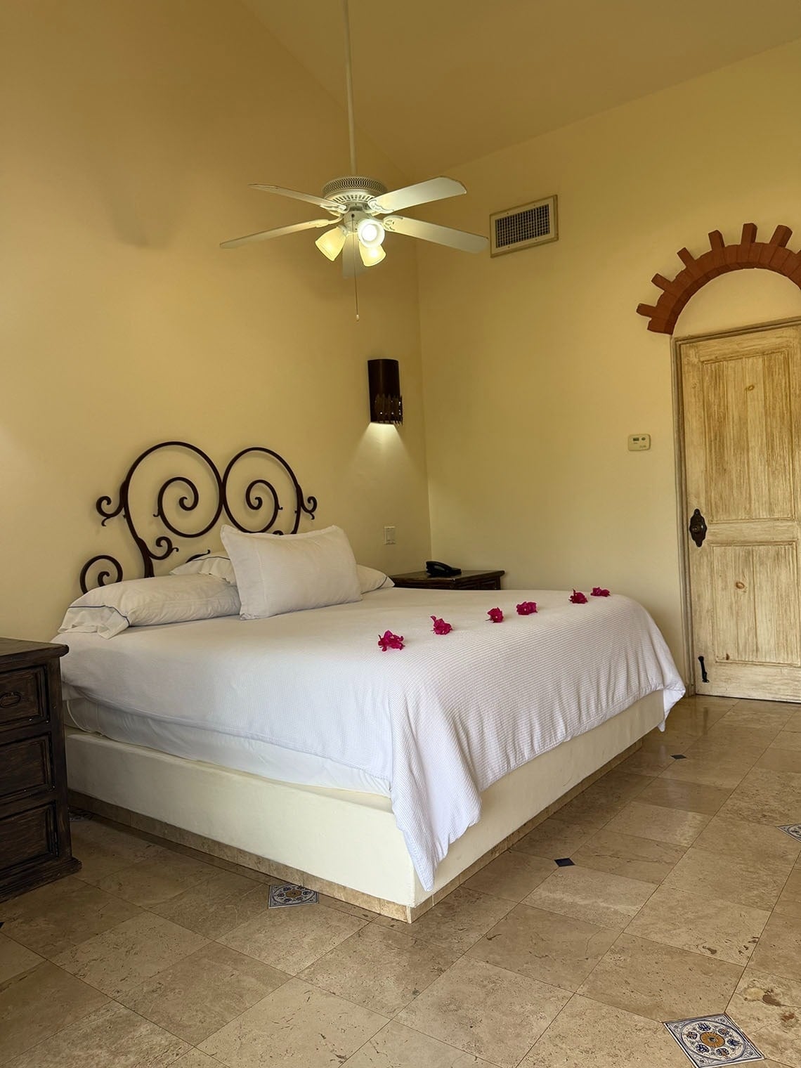 Una habitación de estilo rústico con una cama grande con cabecera de hierro forjado, decorada con flores, un ventilador de techo y suelo de baldosas.