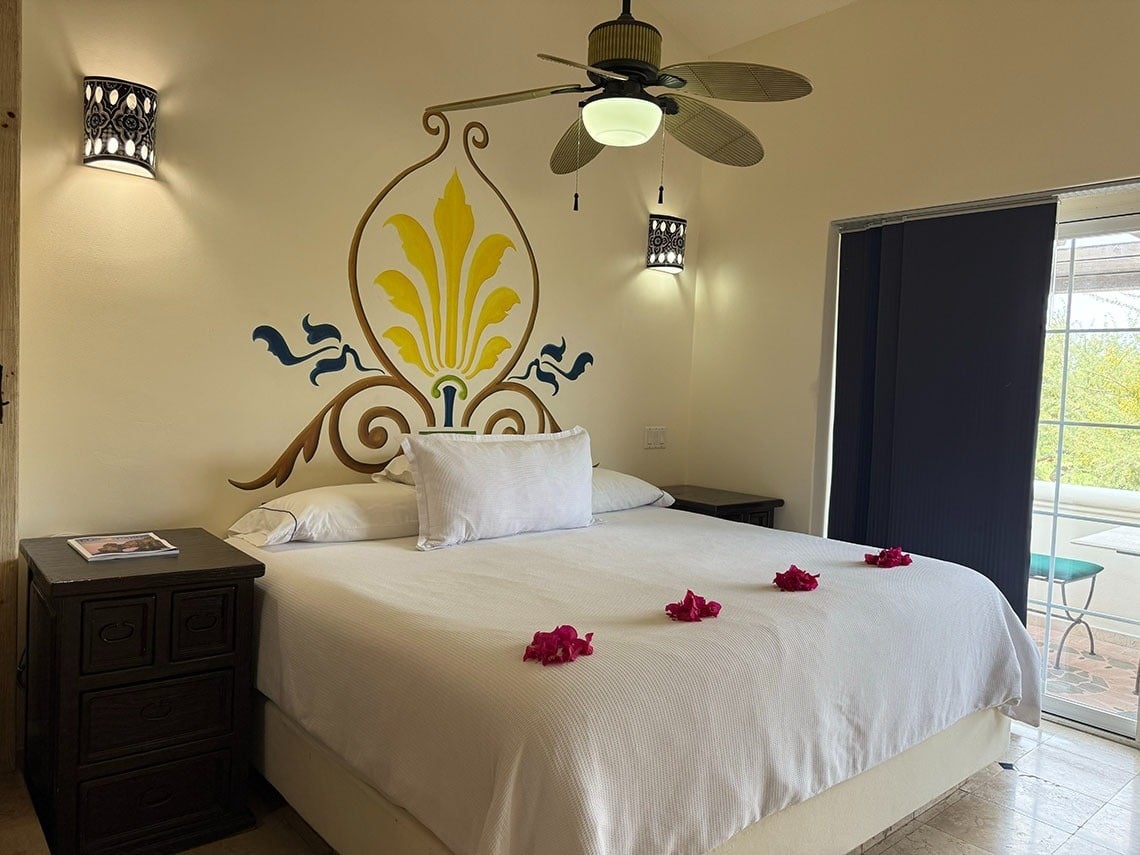 Una habitación de hotel acogedora presenta una cama grande decorada con pétalos, un ventilador de techo, apliques de pared y un vibrante mural en la cabecera, con una puerta que da a un balcón.