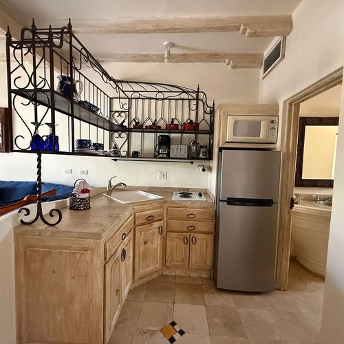 Una cocineta de estilo rústico, con gabinetes de madera, estantería de hierro forjado, electrodomésticos y un sofá azul, con acceso a un baño.