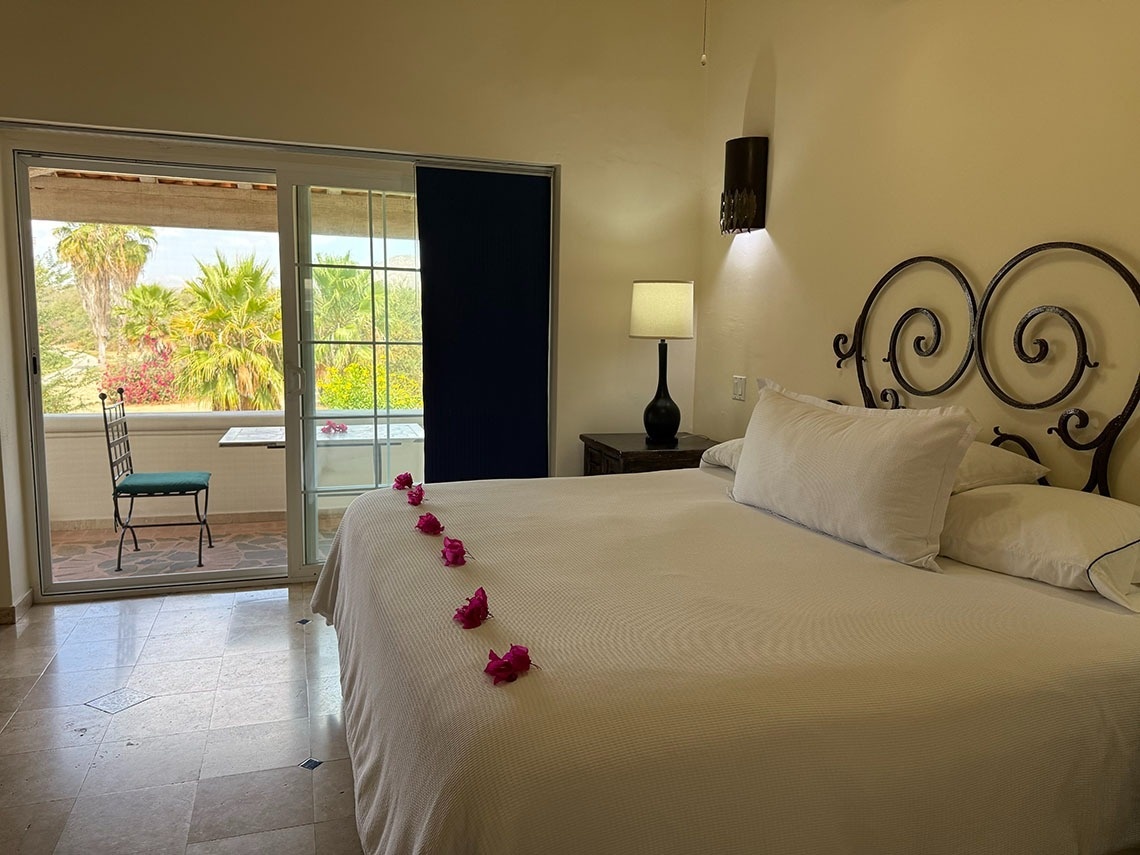 Un dormitorio con cama adornada con flores rojas, una cabecera de hierro forjado y una puerta corrediza que se abre a un balcón con vista a palmeras y exuberante vegetación.