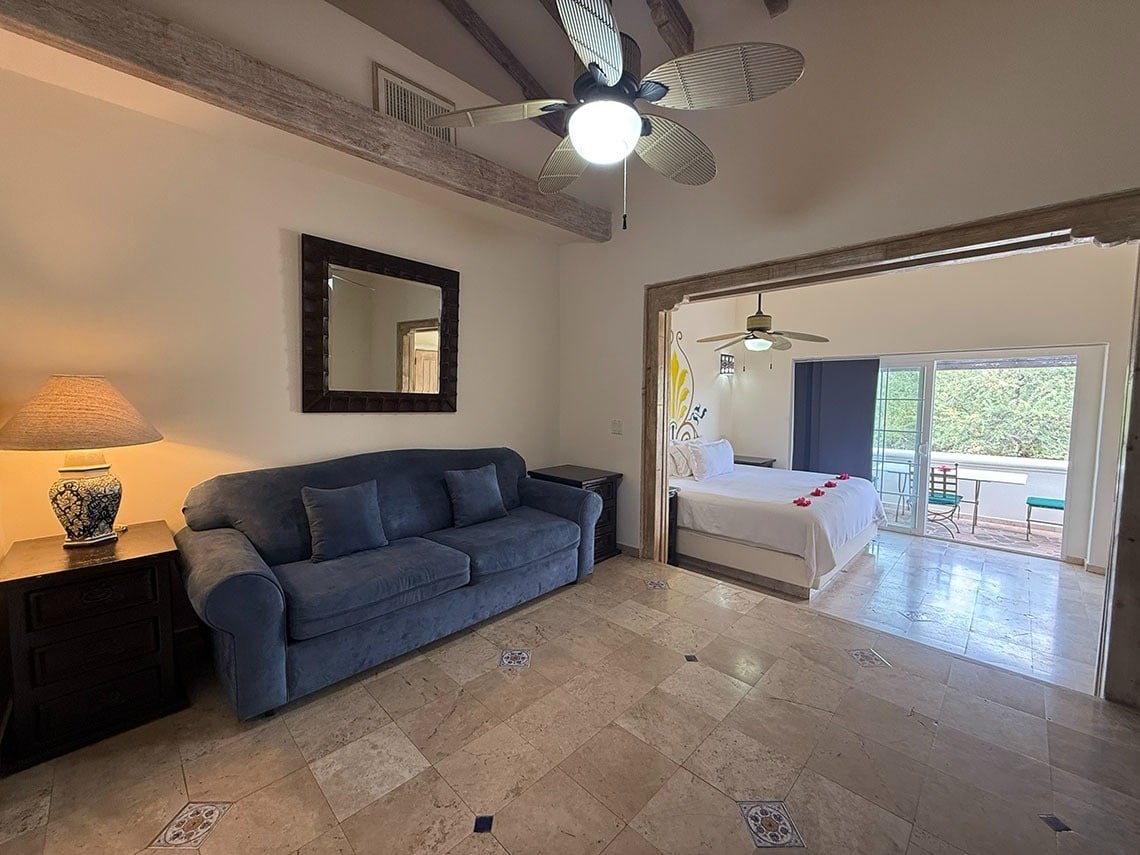 Un amplio espacio interior que incluye una sala de estar con sofá azul y un dormitorio con cama y balcón con vistas a la vegetación, ambos conectados y con suelos de baldosas, vigas de madera y ventiladores de techo.