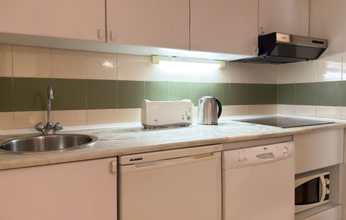 A compact kitchen features a light-colored countertop with a stainless steel sink, toaster, kettle, and electric cooktop, surrounded by white upper and lower cabinets, integrated appliances, and a tiled backsplash with white and green stripes.