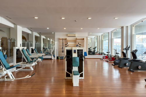 A imagem mostra uma academia espaçosa e bem iluminada com piso de madeira, diversos equipamentos de ginástica, espelhos refletindo o ambiente e grandes janelas.
