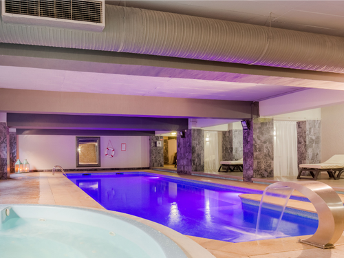 Uma moderna área de spa interior apresenta uma piscina com iluminação azul vibrante, jacuzzi e espreguiçadeiras, oferecendo um ambiente relaxante.
