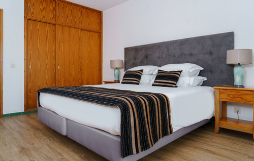A bedroom features a bed with a gray tufted headboard, white bedding, and striped accents, flanked by wooden nightstands with lamps, and a large built-in wooden wardrobe on the left, all set on a light wood floor.