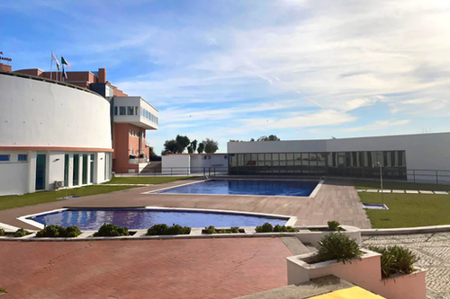 Un complejo moderno presenta dos piscinas al aire libre, rodeadas de edificios blancos y zonas verdes, bajo un cielo azul con nubes dispersas.