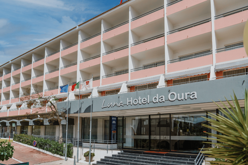 Uma vista externa do Luna Hotel da Oura, um edifício de vários andares com varandas cor-de-rosa e brancas e uma entrada principal em tom cinzento, sob um céu azul.