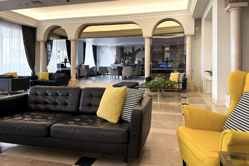 Um lobby sofisticado com sofás de couro preto e detalhes em amarelo vibrante, emoldurado por arcos clássicos que dão para uma área de bar.