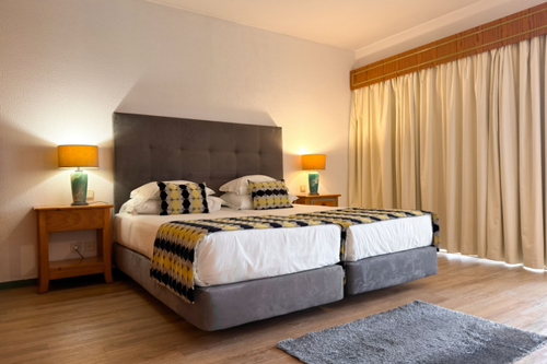 Um quarto de hotel apresenta duas camas com cabeceira cinza, roupa de cama com padrão geométrico amarelo e preto, ladeadas por mesas de cabeceira com abajures, e cortinas claras sobre um piso de madeira.