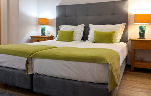 A well-lit hotel room features two single beds with white bedding, lime green throws and accent pillows, a shared gray tufted headboard, and wooden nightstands with illuminated lamps on either side.