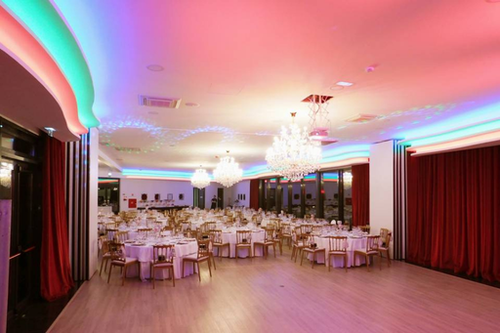 Una amplia y elegante sala de eventos está preparada con mesas redondas vestidas, sillas de madera, grandes lámparas de araña de cristal, cortinas rojas y una vibrante iluminación LED de colores en el techo.