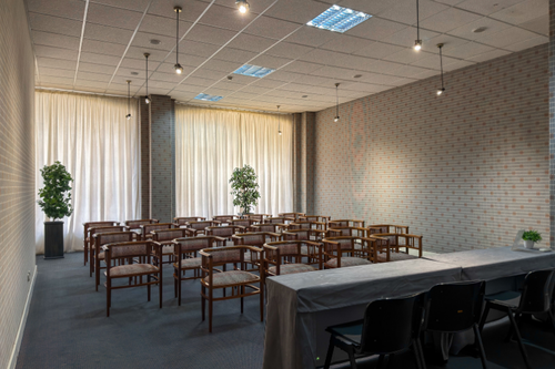 Uma sala de conferências vazia apresenta cadeiras de madeira dispostas em filas, uma mesa coberta com toalha à frente, cortinas claras nas janelas e paredes com padrão de tijolos.