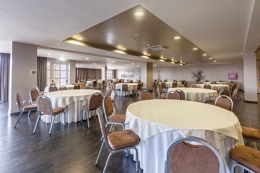 Um salão de eventos espaçoso está montado com muitas mesas redondas cobertas com toalhas de mesa claras e cadeiras.