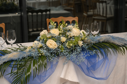A beautifully decorated table features an elaborate floral centerpiece with white roses, blue baby's breath, green foliage, and blue feathery elements, draped with sheer blue fabric and set with wine glasses.
