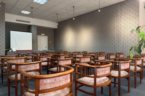 Uma sala de conferências ou reuniões vazia é apresentada, com várias cadeiras de madeira e estofamento estampado dispostas em filas, um telão de projeção e uma mesa na frente, sob luzes suspensas e paredes texturizadas.